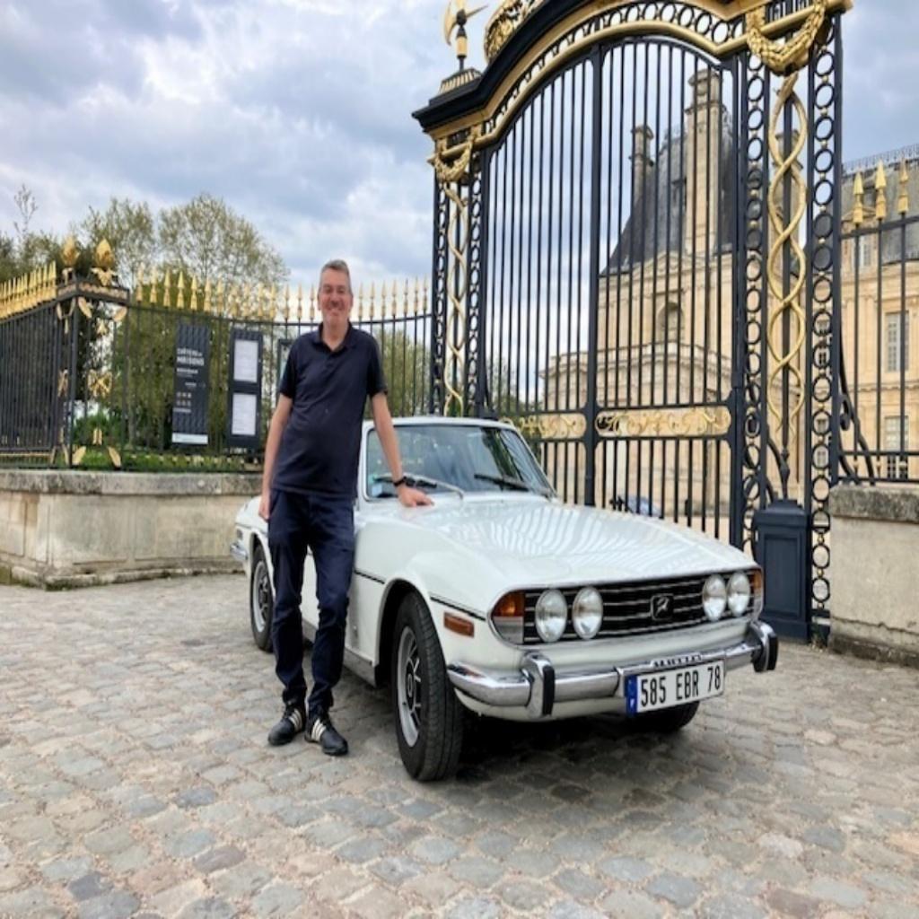 Simon Williams beside his white Triumph Stag at the gates of the Château de Maisons-Laffitte